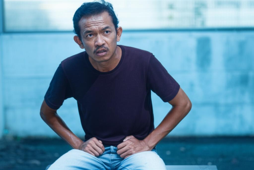An Indonesian man sitting uncomfortably, holding his lower abdomen, looking concerned, with a background suggesting a slightly damp or cool environment.