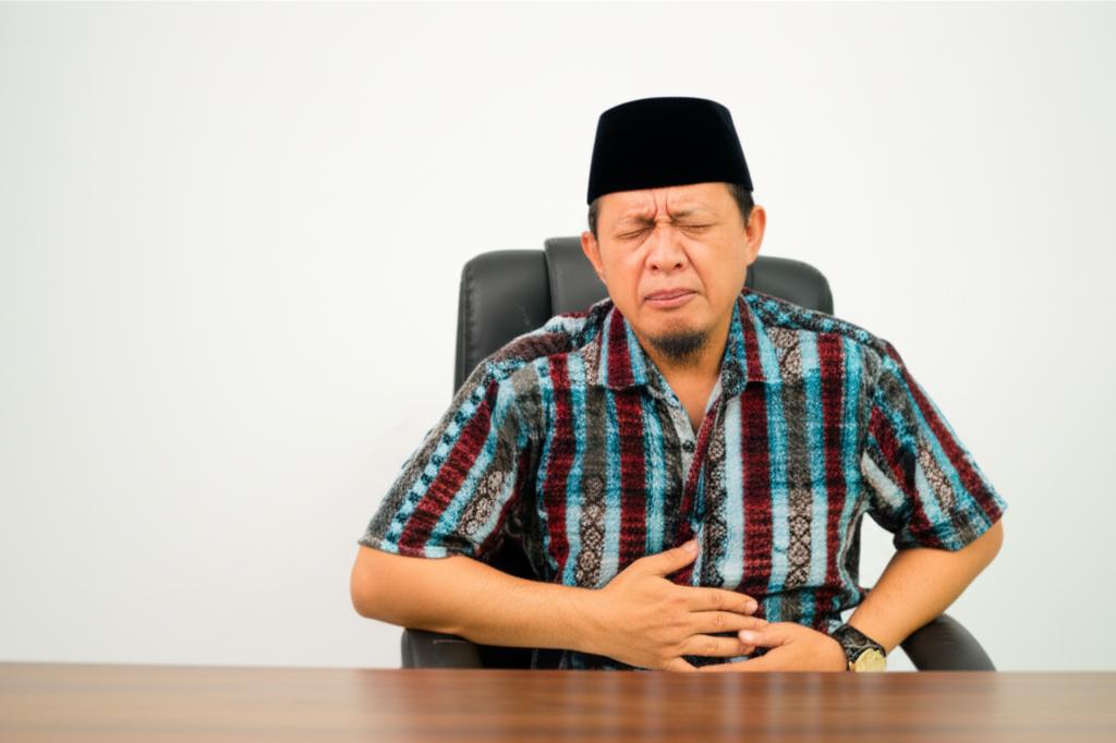 A middle-aged Indonesian man sitting at a desk, looking uncomfortable and holding his lower abdomen.