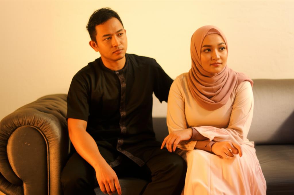 AI prompt: A young Indonesian couple sitting on a sofa looking slightly distant, with the man looking tired and thoughtful. Warm, soft lighting. Realistic photo style.