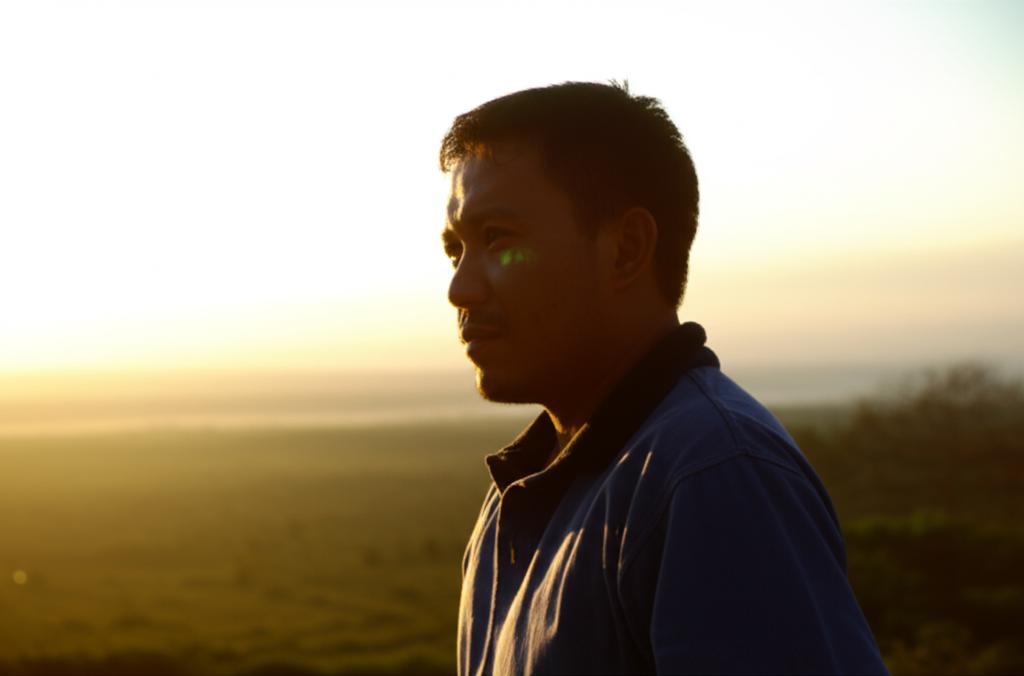 A thoughtful Indonesian man looking out over a serene natural landscape at sunset, contemplating health and well-being, natural light, cinematic